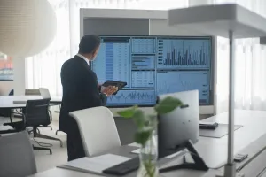 A man wearinga black  suit standing in front of a screen with financial figures on display